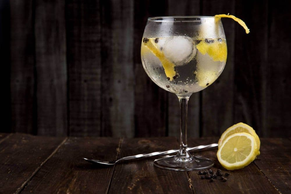 A full cocktail glass with lemon garnish sitting on a dark wooden table with a cocktail spoon and lemon slices, in front of a dark wooden board background.
